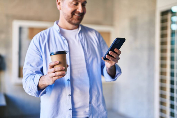 Νεαρός καυκάσιος άνδρας που χρησιμοποιεί smartphone πίνοντας καφέ στο δρόμο - Φωτογραφία, εικόνα