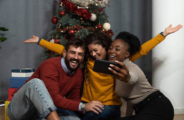 Three young people exchange students roommates and close friends celebrating New Year and Christmas Eve in the apartment with gifts and decorated Christmas Tree. Holidays concept. - Фото, изображение