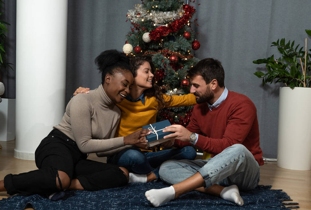 Three young people exchange students roommates and close friends celebrating New Year and Christmas Eve in the apartment with gifts and decorated Christmas Tree. Holidays concept. - Foto, Imagem