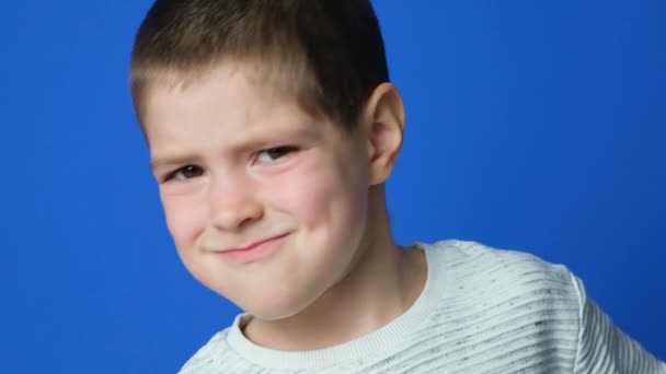 Ujo 5-vuotias poika paheksuu, hymyilee, katsoo kameraan sinisellä taustalla - Materiaali, video