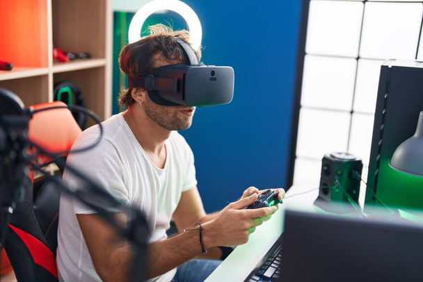 Jeune homme hispanique streamer jouer à un jeu vidéo en utilisant des lunettes de réalité virtuelle et joystick au studio de musique - Photo, image