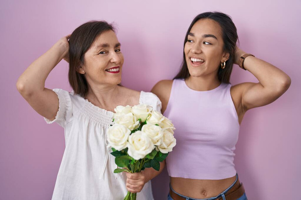 Hispanische Mutter und Tochter halten einen Strauß weißer Blumen in der Hand, lächeln selbstbewusst und berühren das Haar mit erhobener Hand, posieren attraktiv und modisch  - Foto, Bild