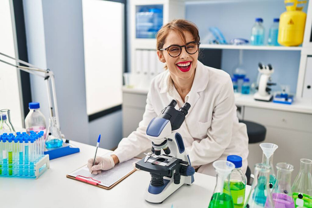 Młoda kaukaska kobieta w mundurze naukowca pisząca na podkładce i mikroskopie uisng w laboratorium - Zdjęcie, obraz