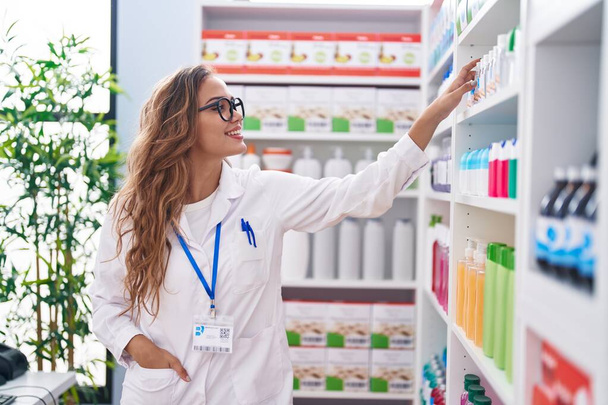 Joven bella mujer hispana farmacéutica sonriendo confiada sosteniendo botella en estanterías en la farmacia - Foto, Imagen