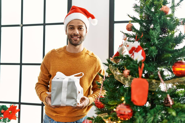 Young arab man smiling happy holding gift standing by christmas tree at home. - 写真・画像