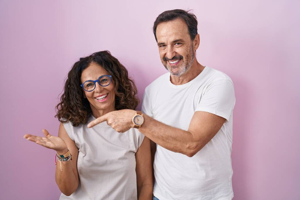 Middle age hispanic couple together over pink background amazed and smiling to the camera while presenting with hand and pointing with finger.  - Фото, зображення