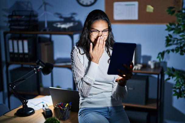 Mujer brasileña joven usando touchpad en la noche trabajando en la oficina riendo y riéndose avergonzado cubriendo la boca con las manos, chismes y concepto de escándalo  - Foto, Imagen