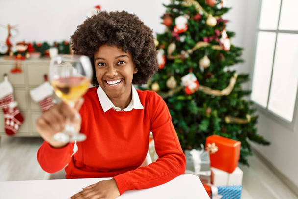 Jeune femme afro-américaine griller avec du vin assis près de l'arbre de Noël à la maison - Photo, image