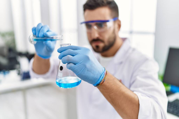 Jovem hispânico vestindo cientista uniforme misturando líquido no laboratório - Foto, Imagem