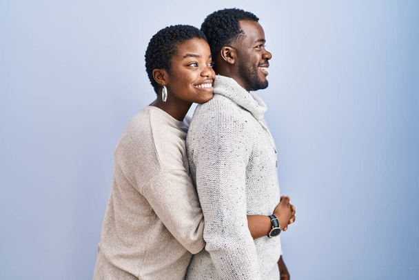 若いアフリカ系アメリカ人のカップルが一緒に側に見て青い背景に立って、自然な顔と自信を持って笑顔でプロファイルポーズをリラックス.  - 写真・画像
