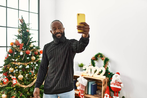 Joven afroamericano hombre hacer selfie por el teléfono inteligente de pie junto al árbol de Navidad en casa - Foto, Imagen