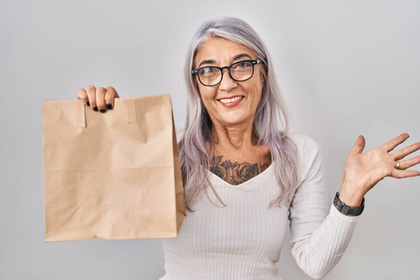 Mujer de mediana edad con pelo gris sosteniendo llevar bolsa de papel celebrando el logro con sonrisa feliz y la expresión ganadora con la mano levantada  - Foto, imagen