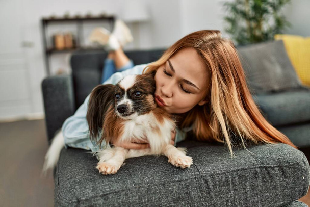 Young caucasian woman lying on sofa hugging and kissing dog at home - Foto, imagen