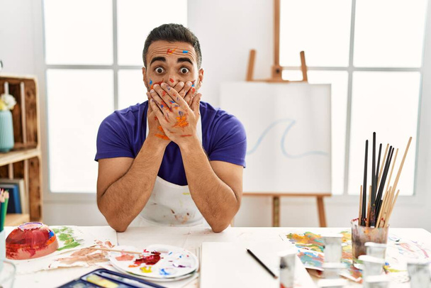 Junger hispanischer Mann mit Bart im Kunstatelier mit gemaltem Gesicht, das den Mund mit den Händen versehentlich verdeckt. Geheimes Konzept.  - Foto, Bild