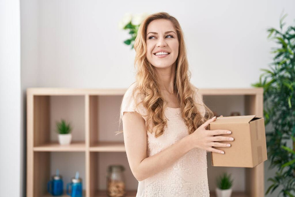 Joven mujer rubia sonriendo confiado sosteniendo paquete en casa - Foto, Imagen