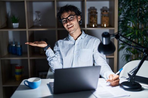 Латиноамериканец, работающий в офисе по ночам, указывая в сторону с открытыми руками ладонями, показывая пространство для копирования, представляя рекламу, улыбающуюся счастливой  - Фото, изображение