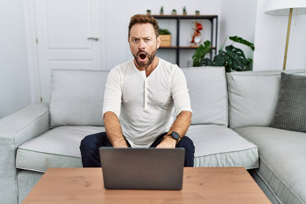 Hombre de mediana edad que utiliza el ordenador portátil en casa en la cara de choque, con aspecto escéptico y sarcástico, sorprendido con la boca abierta  - Foto, imagen
