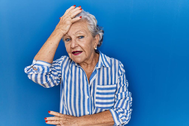 Donna anziana con i capelli grigi in piedi su sfondo blu sorpreso con mano sulla testa per errore, ricordare l'errore. dimenticato, concetto di cattiva memoria.  - Foto, immagini