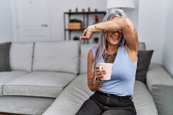 Middelbare leeftijd grijsharige vrouw drinken koffie zitten op de bank thuis glimlachend vrolijk spelen gluren een boo met handen tonen gezicht. verrast en verlaten  - Foto, afbeelding