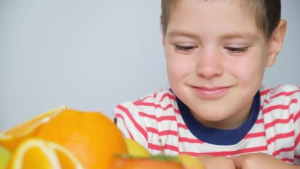 Um menino bonito de 5 anos sorri e olha para a fruta laranja em um fundo branco. - Filmagem, Vídeo
