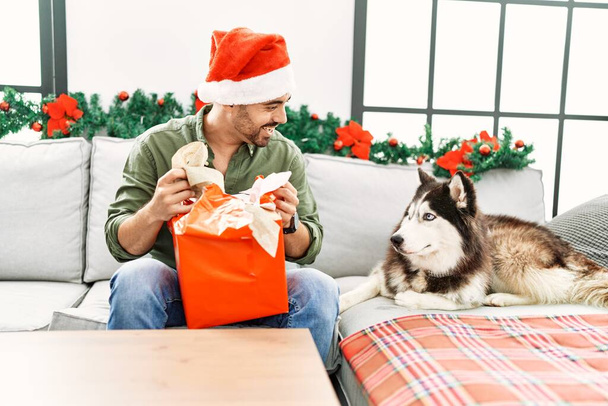 Молодой латиноамериканец раскладывает подарок сидя на диване с собакой под рождественским декором дома - Фото, изображение