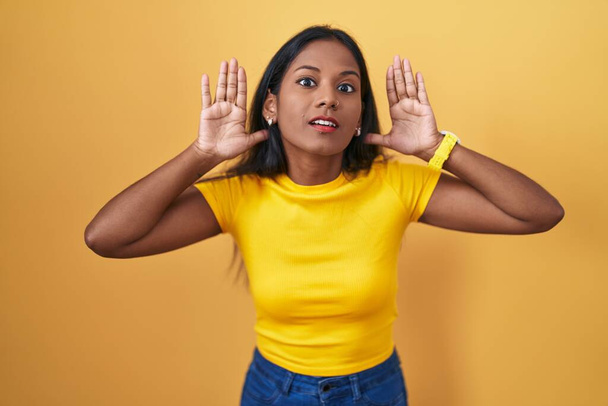 Mujer india joven de pie sobre el fondo amarillo tratando de escuchar ambas manos en el gesto de la oreja, curioso por chismes. problema auditivo, sordo  - Foto, imagen