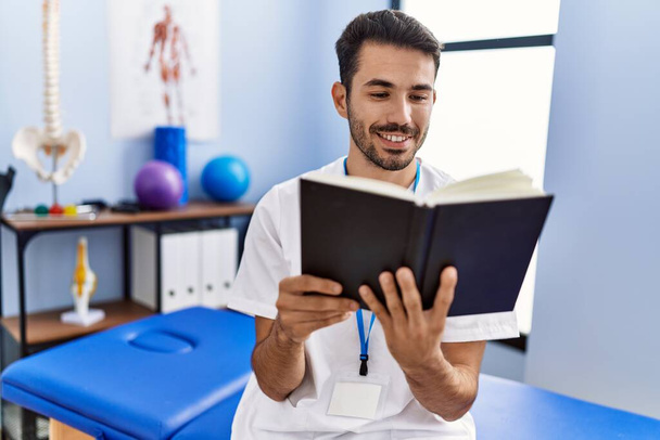 Young hispanic man wearing physiotherapist uniform reading book at rehab clinic - Fotó, kép