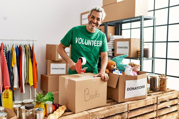 Middle age grey-haired man volunteer smiling confident packing books cardboard box at charity center - Photo, Image
