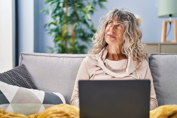 Mujer de mediana edad utilizando el ordenador portátil sentado en el sofá en casa - Foto, imagen