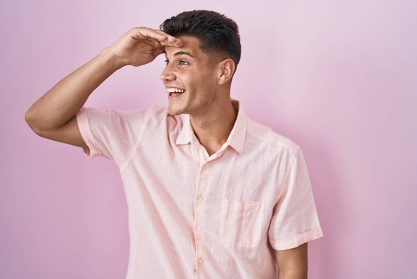 Joven hombre hispano de pie sobre fondo rosa muy feliz y sonriente mirando lejos con la mano sobre la cabeza. concepto de búsqueda.  - Foto, imagen