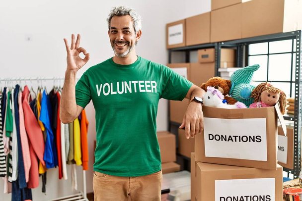 Middle age hispanic man wearing volunteer t shirt at donations stand smiling positive doing ok sign with hand and fingers. successful expression.  - Photo, Image