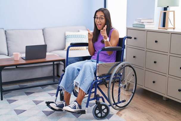 Mulher hispânica jovem sentada em cadeira de rodas em casa apontando os dedos para a câmera com rosto feliz e engraçado. boa energia e vibrações.  - Foto, Imagem