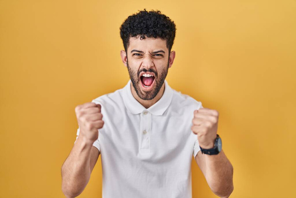 Arab man standing over yellow background angry and mad raising fists frustrated and furious while shouting with anger. rage and aggressive concept.  - Foto, afbeelding