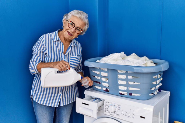 Μεγαλύτερη γκρίζα μαλλιά γυναίκα χαμογελά αυτοπεποίθηση ρίχνει απορρυπαντικό στο πλυντήριο στο δωμάτιο πλυντηρίων - Φωτογραφία, εικόνα