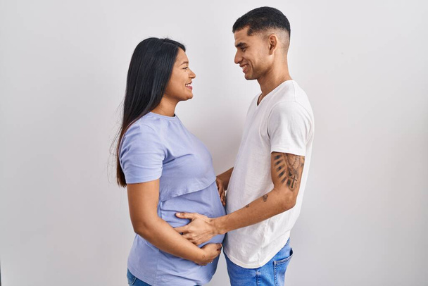 Junges hispanisches Paar erwartet ein Baby, das über dem Hintergrund steht und zur Seite schaut, entspannte Profil-Pose mit natürlichem Gesicht und selbstbewusstem Lächeln.  - Foto, Bild