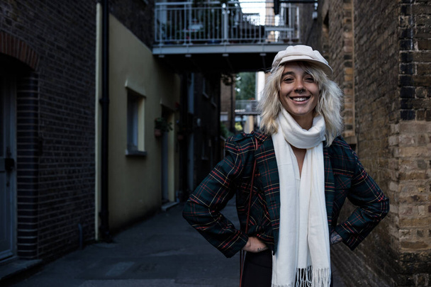 Улыбающаяся и счастливая женщина-модель смотрит в камеру на узкой улице. На ней белая кепка и шарф.. - Фото, изображение