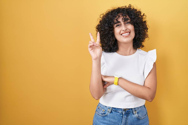 Young middle east woman standing over yellow background smiling with happy face winking at the camera doing victory sign. number two.  - Photo, Image