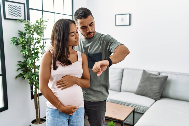 Giovane coppia interrazziale in attesa di un bambino, toccando pancia incinta controllando il tempo su orologio da polso, rilassato e fiducioso  - Foto, immagini