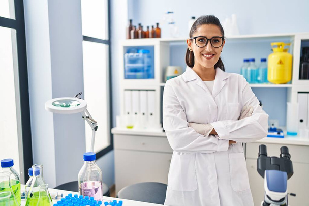 Młoda Latynoska kobieta w mundurze naukowca stojąca z rękoma skrzyżowanymi w laboratorium - Zdjęcie, obraz
