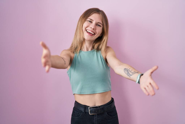 Blonde caucasian woman standing over pink background looking at the camera smiling with open arms for hug. cheerful expression embracing happiness.  - Photo, Image