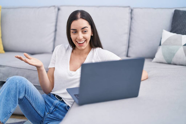 Jeune femme brune utilisant un ordinateur portable à la maison célébrant la réalisation avec un sourire heureux et l'expression gagnante avec la main levée  - Photo, image