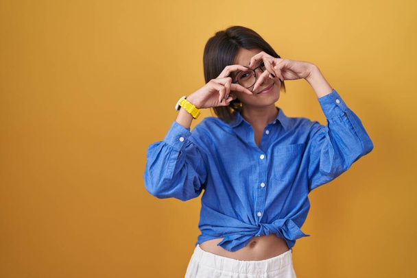 Jeune fille debout sur fond jaune faisant forme de coeur avec la main et les doigts souriant en regardant à travers signe  - Photo, image