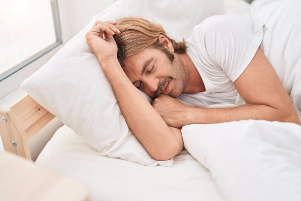 Young blond man lying on bed sleeping at bedroom - Фото, зображення