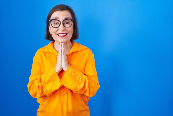 Orta yaşlı İspanyol bir kadın mavi arka planda gözlük takıyor. Elleriyle birlikte dua ediyor. Affedilmek için dua ediyor..  - Fotoğraf, Görsel