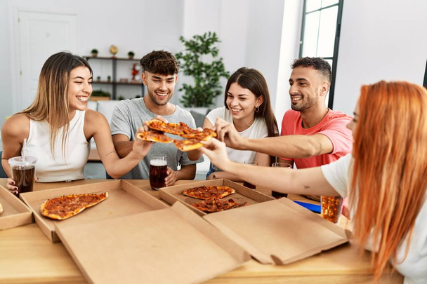 Gruppe junger Freunde lächelt glücklich beim Essen italienischer Pizza zu Hause auf dem Tisch sitzend. - Foto, Bild