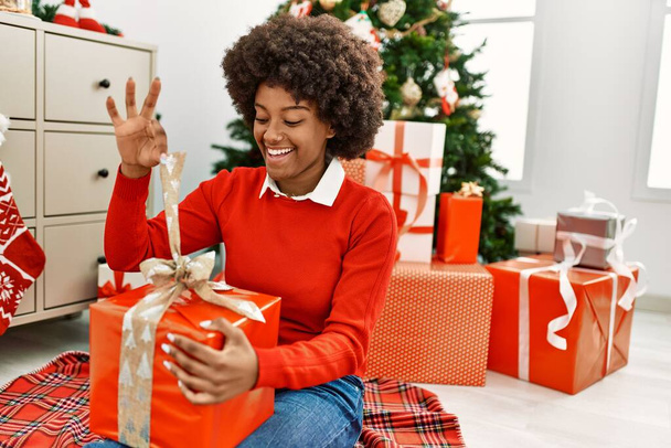 Young african american woman unboxing gift sitting by christmas tree at home - 写真・画像