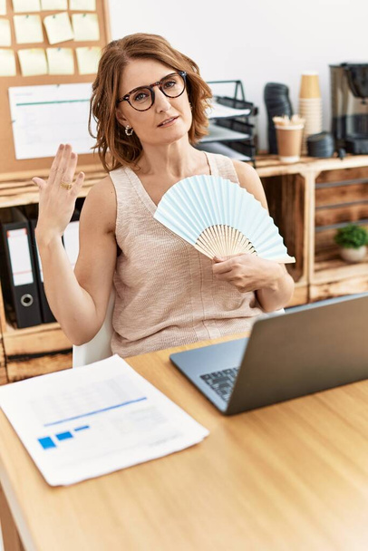 Middle age hispanic woman using handfan working at office - Foto, Imagen