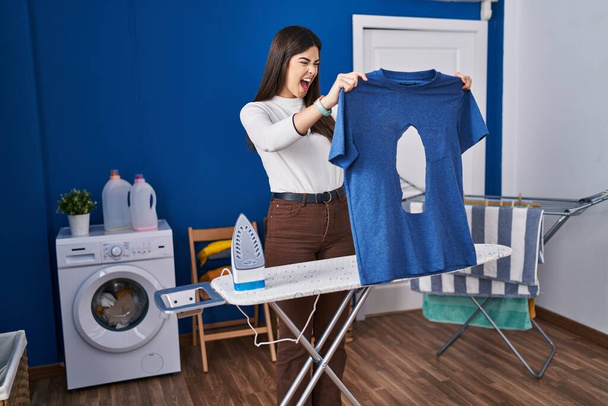 Young brunette woman ironing holding burned iron shirt at laundry room angry and mad screaming frustrated and furious, shouting with anger. rage and aggressive concept.  - Photo, Image