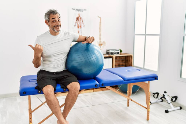 Hombre hispano de mediana edad en la clínica de recuperación del dolor sosteniendo la pelota de pilates apuntando a la espalda con la mano y los pulgares hacia arriba, sonriendo confiado  - Foto, Imagen