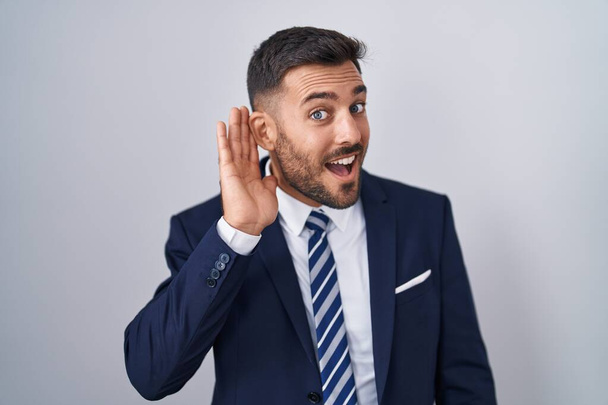 Hombre hispano guapo vistiendo traje y corbata sonriendo con la mano sobre la oreja escuchando un rumor o chismes. concepto de sordera.  - Foto, imagen